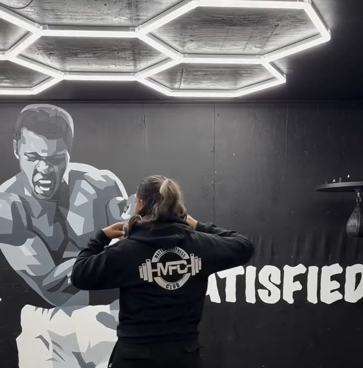Espalda de la sudadera negra MFC con el logo blanco de Mattawa Fitness Club, frente al mural de Never Satisfied